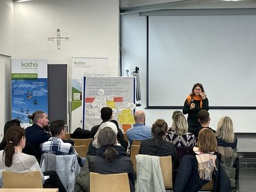 Das Foto zeigt einen Seminarraum, vorne steht eine weiblich gelesene Person, die einen Vortrag hält. Die Teilnehmer_innen sitzen auf Stühlen und sind von hinten aufgenommen, sie folgen dem Vortrag.