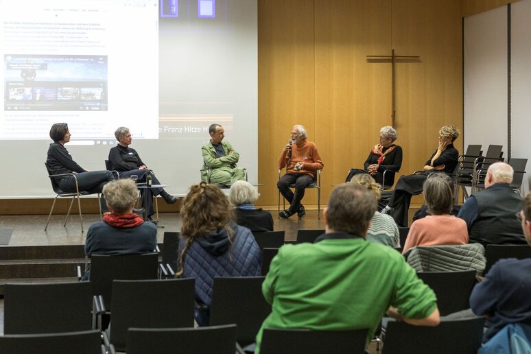 Sechs Personen sitzen in einem Halbkreis vor Publikum auf der Bühne. Ein Mann im orangen Pullover hat das Mikrofon und spricht zu den Anderen.