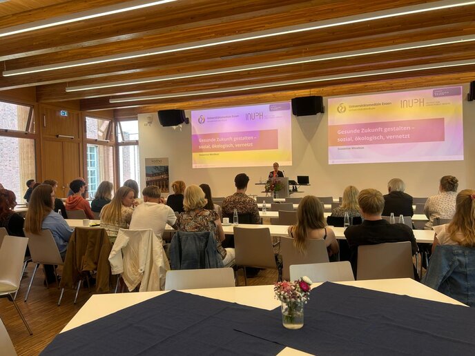 Prof.in Dr.in Susanne Moebus hielt einen Vortrag über die Verknüpfung zwischen Umwelt und Gesundheit. Sie steht auf der Bühne und referiert. Im Vordergrund hören die Zuschauer_innen zu.