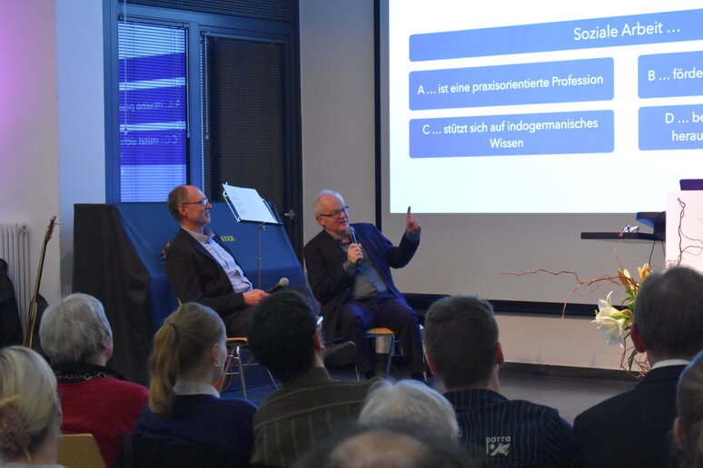 Jörg Baur und Christof Stock sitzen auf der Bühne und müssen eine Frage beantworten. Die Frage und vier Antwortmöglichkeiten stehen auf der Folie. Die