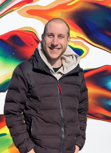 Der Student Benjamin Karenke steht vor einer bunten Wand und lächelt in die Kamera.
