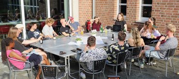 Ca. 15 Studierende sitzen an einem großen Tisch auf einer Terrasse und blicken in Richtung der Dozentin, die etwas auf einem Laptop präsentiert..