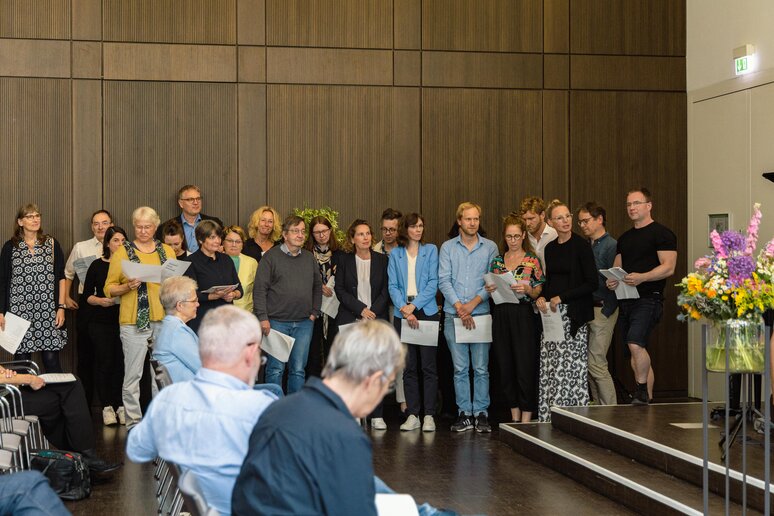Kolleg_innen singen in einem Chor zusammen; begleitet werden sie von Klavier und Gitarre. Die Geehrten hören gespannt zu.