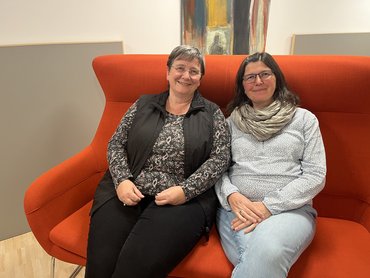 Christine Geier (links) und Anne Würth sitzen auf einem roten Sofa nebeneinander und lächeln freundlich in die Kamera.
