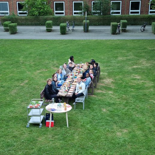 Studierende sitzen draußen auf einer grünen Wiese an einem langen Tisch mit Essen vor sich.