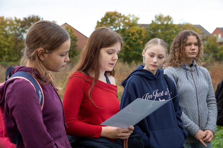Vier Schülerinnen stehen nebeneinander auf einer Wiese. Eine liest einen laminierten Zeitungsartikel vor.