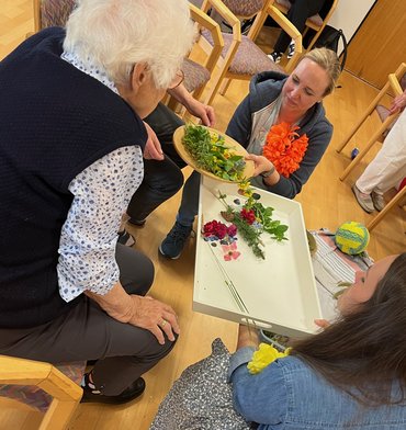 Eine junge Frau reicht einer Seniorin einen Teller mit grünen Zweigen. Die Seniorin ist von hinten, sitzend auf einem Stuhl zu sehen, die junge Frau von vorne.