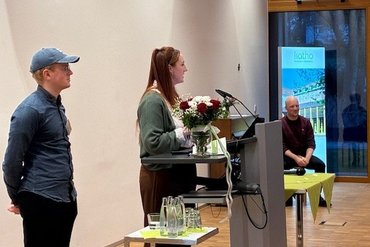 Zwei Studierende stehen am Rednerpult und gratulieren den Absolvent_innen