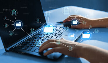 Im Vordergrund ist virtuell eine Dateistruktur abgebildet. Im Hintergrund sitzt eine Person an einem Tisch mit ihrem Laptop.