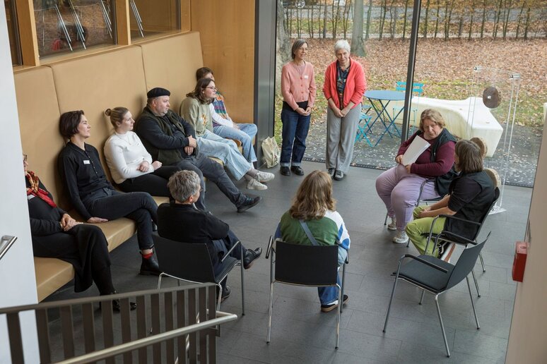 Eine Gruppe Menschen sitzt im Kreis zusammen. Eine Hälfte auf einer Bank, die anderen auf Stühlen. Alle Teilnehmenden schauen in die Richtung einer jungen Frau, die auch mit im Kreis sitzt.