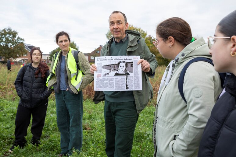 Ein Mann hält einen Zeitungartikel in die Kamera. Um ihn herum stehen vier Menschen. Einige Schüler:innen laufen im Hintergund herum.