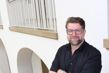 Dr. Jens Gründler steht für ein Portraitfoto vor einem weißen Gebäude.
