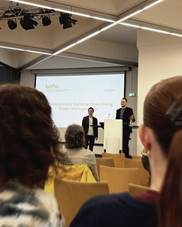 Zwei Männer stehen am bzw. neben dem Rednerpult, einer redet, der andere hört zu.