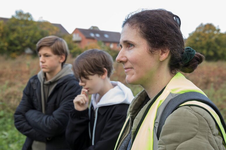 Eine Frau und zwei Schüler stehen auf einer Wiese. Alle schauen aus dem Bild raus, nicht in die Kamera.