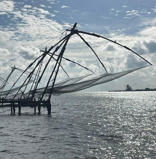 Chinesische Fischernetze an hölzernen Gestellen über dem Meer; glitzerndes Wasser und wolkiger, leicht bewölkter Himmel im Hintergrund.