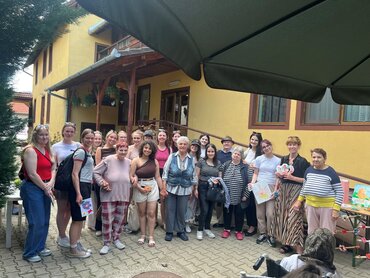 Eine Gruppe von Menschen steht in drei Reihen hintereinander vor einem gelben Haus, mit einem Geländer an der rechten Seite.