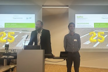 Prof. Dr. Ulrich Frischknecht und Dr. Felix Müller am Redepult. Im Hintergrund ist das Titelbild der Fachtagung mit einer goldenen 25 projiziert.