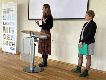 Dr.in des. Anja Schmid-Engbrodt und Dr.in Judith Schmidt stehen auf der Bühne und halten eine Präsentation, die hinter ihnen an die Wand projiziert wird. Links neben ihnen steht das Plakat der Ausstellung.