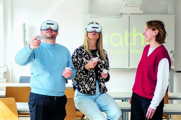 Zwei Personen in einem Seminarraum tragen VR-Brillen, eine weitere beobachtet die beiden.uen umher