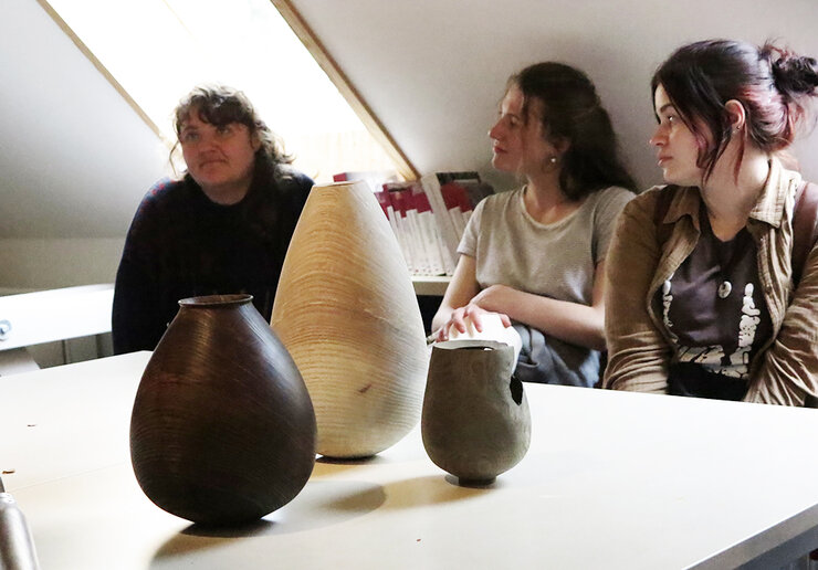 Drei Frauen sitzen um einen kleinen Tisch herum. Im Vordergrund auf dem Tisch stehen drei große leere Vasen.