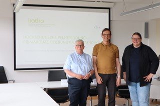Drei katho-Lehrende stehen vor einer Leinwand mit einer Präsentation.