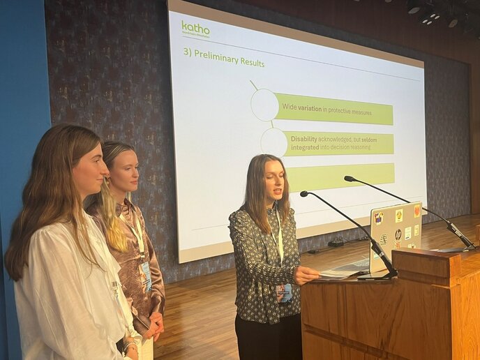 Drei Frauen stehen an einem Rednerpult auf einer Bühne und halten eine Präsentation. Eine von ihnen spricht ins Mikrofon, während die anderen zuhören. Auf der Leinwand hinter ihnen sind die Worte "Preliminary Results" und einige Bulletpoints zu sehen, die Schutzmaßnahmen und Behinderungen thematisieren.