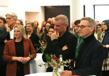 Andreas Reiners steht gemeinsam mit dem Dekan und einer weiteren Frau am Stehtisch und lauscht dem Vortrag für ihn.