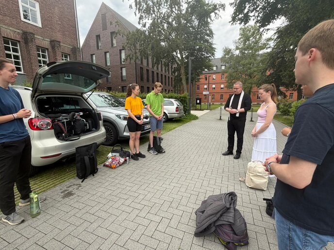 Junge Menschen stehen mit einem Priester zusammen auf einem Parkplatz und falten de Hände zum Gebet.