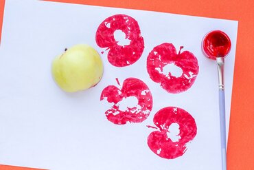 Auf einem weißen Papier sind vier rote Abdrücke eines Apfels gestempelt. Daneben liegen ein hellgrüner Apfel und ein Pinsel mit roter Farbe.