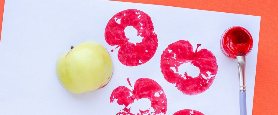 Auf einem weißen Papier sind vier rote Abdrücke eines Apfels gestempelt. Daneben liegen ein hellgrüner Apfel und ein Pinsel mit roter Farbe.