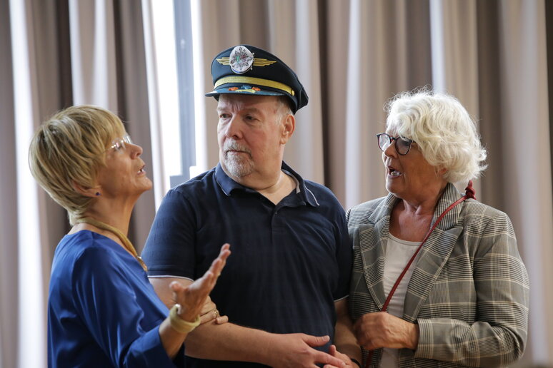 Drei ältere Menschen stehen vor einem Vorhang. Eine Frau in blauem Oberteil spricht gestikulierend mit einem Mann in dunkler Uniform und Schirmmütze, eine weitere Frau im karierten Blazer steht neben ihm.