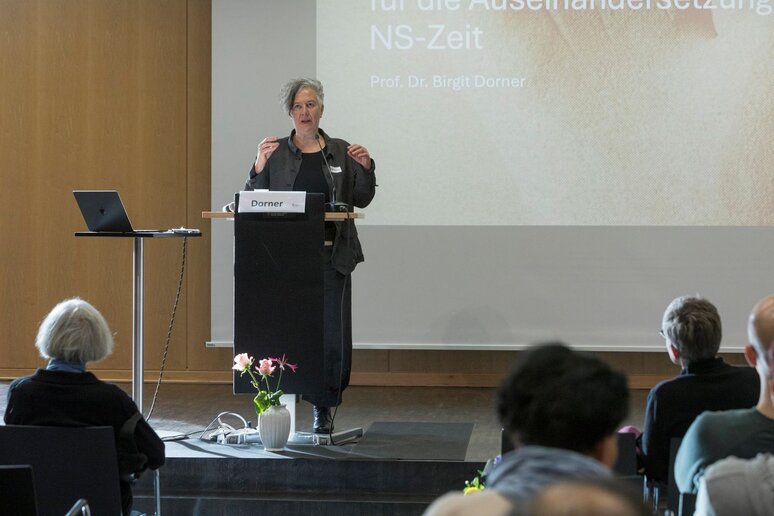 Eine Frau steht am Rednerpult. Im Hintergrund ist eine Präsentation über die NS-Zeit von Prof. Dr. Birgit Dorner.