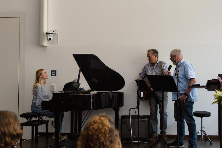 Drei Kolleg_innen musizieren gemeinsam auf der Bühne: Am Klavier, an der Gitarre und mit Gesang