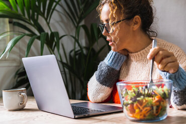 Eine Person, die mit ihrem Laptop am Schreibtisch sitzt und dabei isst.