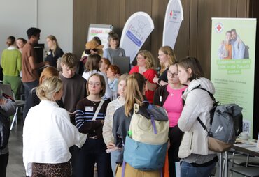 Junge Menschen stehen in Gruppen beisammen und unterhalten sich. Im Hintergrund stehen roll-ups und beachflags, die für Praxiseinrichtungen werben.