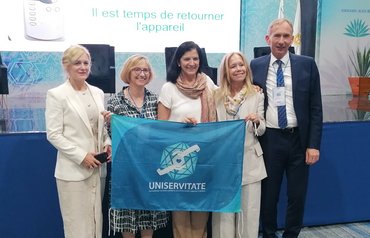 Fünf Personen halten eine blaue Fahne mit der Aufschrift "Uniservitate" und lächeln in eine Richtung.