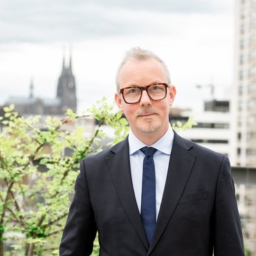 Prorektor Michael Isfort steht auf dem Balkon und er lächelt in die Kamera. Im Hintergrund ist der Dom zu sehen.