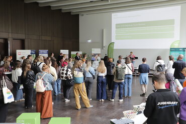 Viele junge Menschen stehen in einem großen Saal mit Blick auf die Vortragende vorne auf der Bühne.