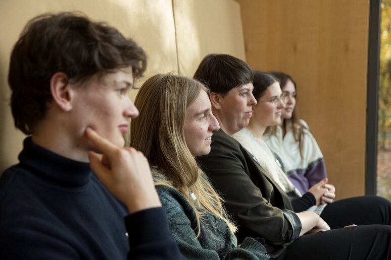 Fünf Menschen sitzen auf einer Bank und schauen nach vorne. Sie sind von der Seite fotografiert, sodass die letzte Person unscharf erscheint.