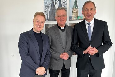 Dompropst Monsignore Joachim Göbel mit seinem Stellvertreter Thomas Pitsch und Geschäftsführer Bernward Robrecht stehen vor einer weißen Wand und lächeln in die Kamera.