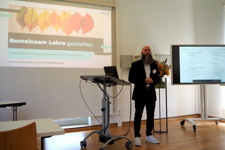 Prof. Arnold steht im Seminarraum neben dem Redepult. Links hinter ihm ist eine Präsentation zu sehen mit dem Titel "Gemeinsam Lehre gestalten".