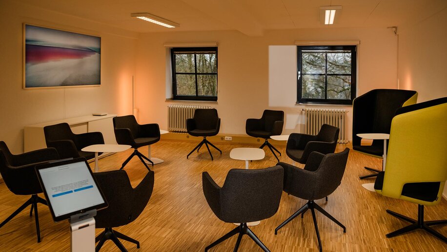 In einem Raum mit zwei Fenstern, Holzboden und warmer Lichtoptik stehen mehrere moderne Drehstühle im Kreis. Am rechten Bildrand stehen zwei weitere Drehstühle mit Akustikschutz. Im Vordergrund steht eine Bedienkonsole, ander Wand links ein großes digitales Display.