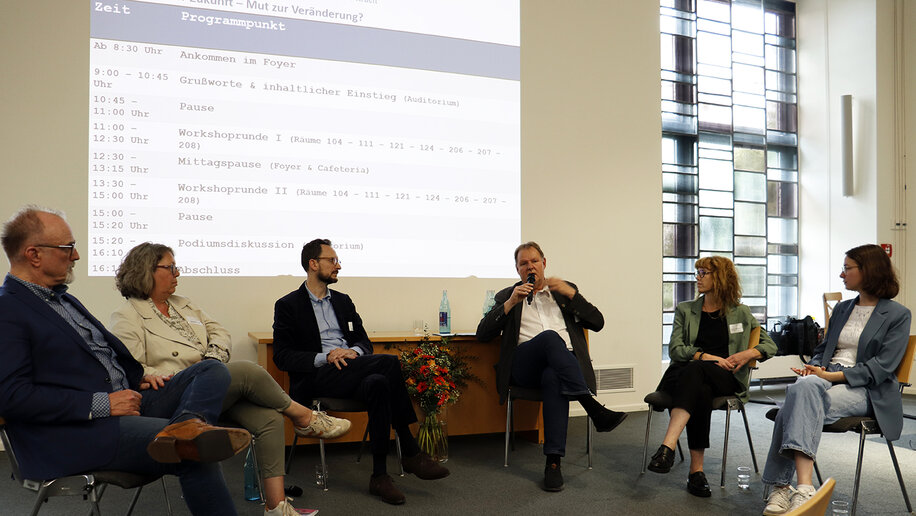 Drei Männer und drei Frauen sitzen in einem Stuhlhalbkreis auf einem Podium. Ein Mann spricht in ein Mikrofon, die anderen wenden sich ihm zu.