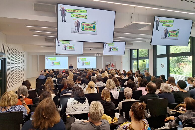 Der Kölner Audimax ist gut gefüllt mit über 200 Menschen. Auf den Monitoren erscheint ein Bild.