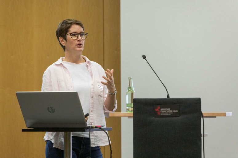 Eine Frau mit kurzen braunen Haaren steht am Rednerpult. Sie trägt weiße Kleidung und eine schwarze Brille.