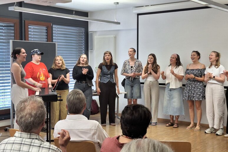 M;ehrere Personen stehen in einem Halbkreis und singen, einige von ihnen klatschen dabei. Neben ihnen spielt eine Frau Keyboard.