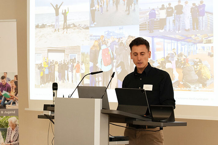 Promovend Lukas Bitzer am Redepult. Hinter ihm eine Präsentation mit Fotos, die Menschen zeigen.