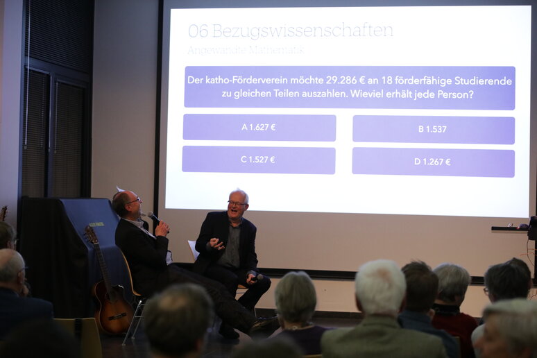 Blick auf die Bühne, im Hintergrund eine Folie mit einer Frage und vier Antwortmöglichkeiten, davor sitzen die beiden Professoren 