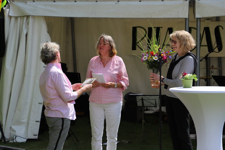 Zwei Frauen überreichen Frau Paß Blumen und eine Karte.