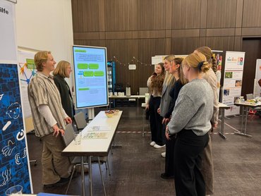 2 Personen stehen hinter einem Tisch; vier Personen vor dem Tisch hören der Vorstellung des Projekts interessiert zu.